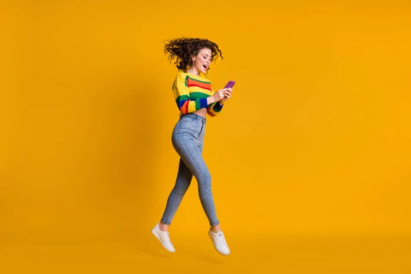 Foto de perfil em tamanho completo de alegre agradável ondulado cabelo marrom menina salto escrever mensagem telefone usar camisola jeans isolado no fundo amarelo — Fotografia de Stock