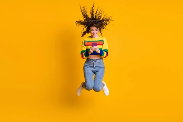 Повнорозмірна фотографія позитивного симпатичного коричневого довгого волосся дівчина стрибає подивитися телефонний одяг пуловер джинси кросівки ізольовані на жовтому фоні — стокове фото
