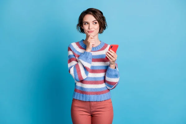 Foto de niña bonita mantenga teléfono celular dedo barbilla mirada lado espacio vacío desgaste a rayas jersey pantalones rojos aislados color azul fondo — Foto de Stock