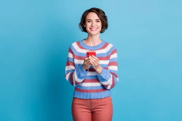 Фото красивої молодої леді тримає телефон блискуча біла посмішка носить барвистий смугастий пуловер червоних штанів ізольований синій колір фону — стокове фото