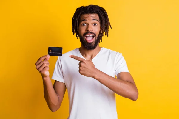 Foto van zwarte stijlvolle man dreadlocks directe wijsvinger houden credit card open mond dragen witte t-shirt geïsoleerde gele kleur achtergrond — Stockfoto