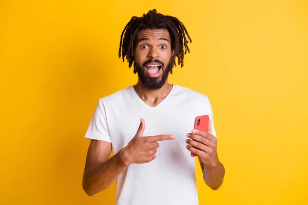 Foto van grappige verbaasd donkere huid guy hold telefoon regisseren vinger scherm apparaat dragen t-shirt geïsoleerde gele kleur achtergrond — Stockfoto