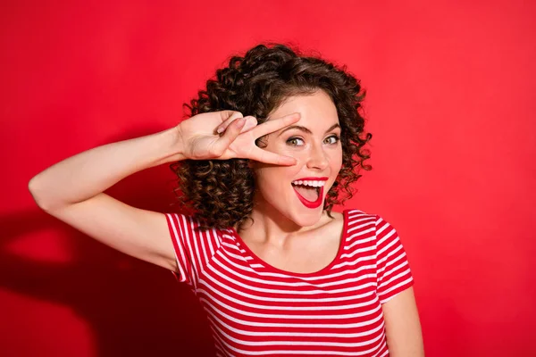 Foto de dulce mujer joven ondulada divertida vestida a rayas que muestra el ojo de signo v aislado fondo de color rojo — Foto de Stock