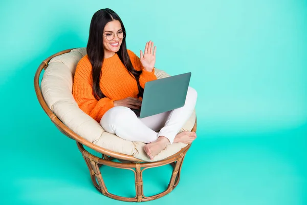 Фото веселой девушки сидеть кресло держать нетбук волна ладони носить очки оранжевый пуловер изолированный бирюзовый цвет фона — стоковое фото