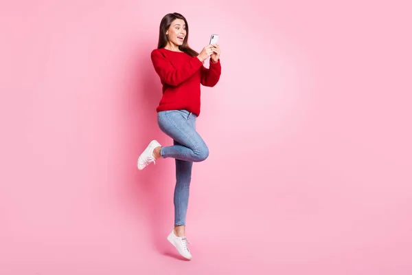 Volledige lengte foto van schattig meisje springen opgewonden scherm hold telefoon dragen rode trui jeans sneakers geïsoleerde roze kleur achtergrond — Stockfoto