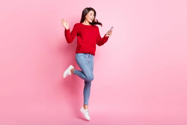 Pleine longueur photo de jolie fille agitant écran main tenir téléphone portable saut porter rouge pull jeans baskets isolé couleur rose fond — Photo