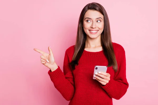 Fénykép a szép lány rendezés mutatóujj nézd oldalon üres tér tartsa mobiltelefon fényes mosoly kopás piros pulóver elszigetelt rózsaszín szín háttér — Stock Fotó