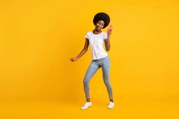 Full length body size photo of beautiful young girl with black skin listening music dancing holding smartphone isolated on bright yellow color background — Stock Photo, Image