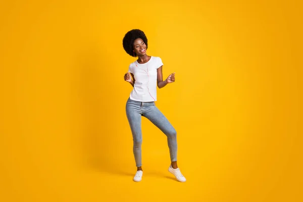 Pełna długość ciała rozmiar zdjęcie uśmiechniętej dziewczyny z czarną skórą słuchanie muzyki taniec trzymając ręce w górze na jasnożółtym tle — Zdjęcie stockowe