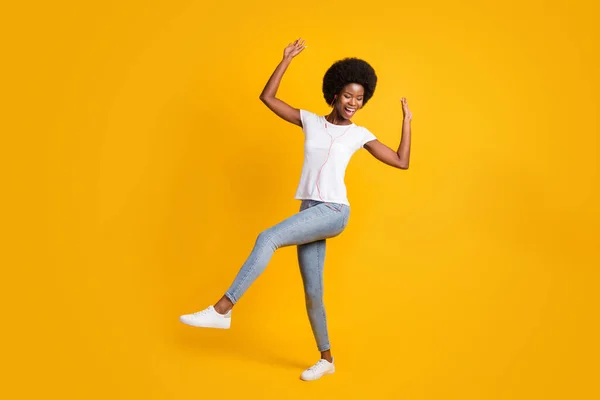 Tamanho total do corpo foto de hipster feminino com pele preta ouvindo música favorita dançando com fones de ouvido laranja sorrindo isolado no fundo de cor amarelo brilhante — Fotografia de Stock