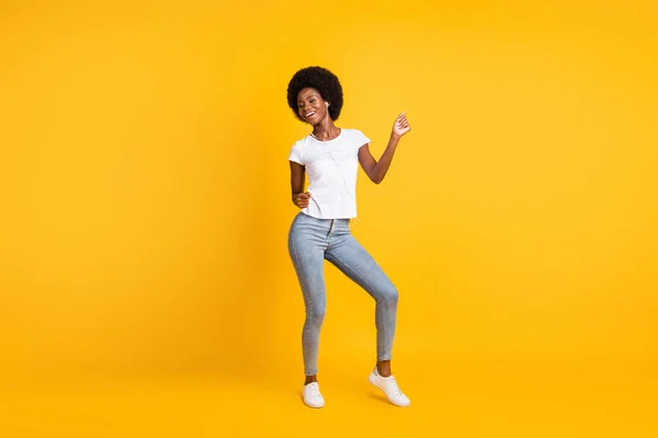 Foto de cuerpo de cuerpo entero de una chica bastante activa con piel negra escuchando música bailando con auriculares usando pantalones vaqueros sonriendo aislados sobre un fondo de color amarillo brillante — Foto de Stock