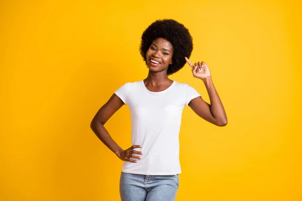 Foto retrato de una joven afroamericana apuntando con el dedo a su cabello afro sonriendo una mano en la cintura usando una camiseta blanca aislada sobre un fondo de color amarillo vivo — Foto de Stock