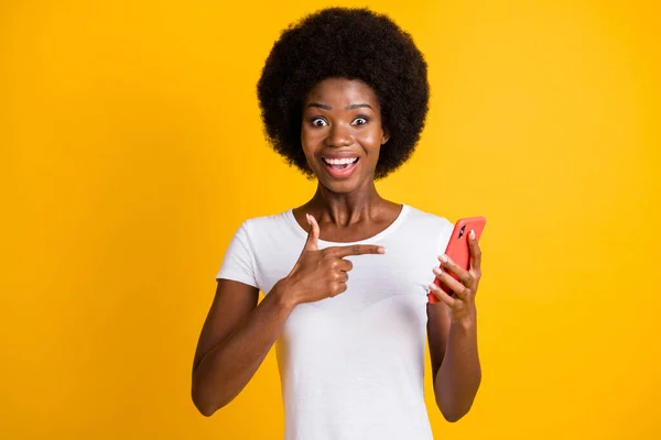 Foto portrét překvapené ženy ukazující prstem na telefon v ruce s otevřenými ústy na sobě ležérní bílé tričko izolované na jasně žlutém pozadí — Stock fotografie