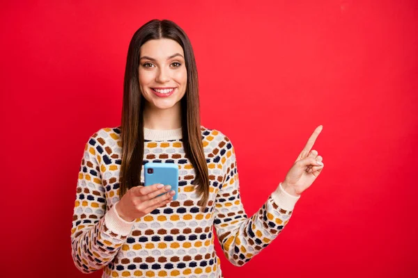 Retrato de otimista tipo menina agradável ponto de telefone espaço vazio desgaste camisola colorida isolado no fundo cor vermelha — Fotografia de Stock