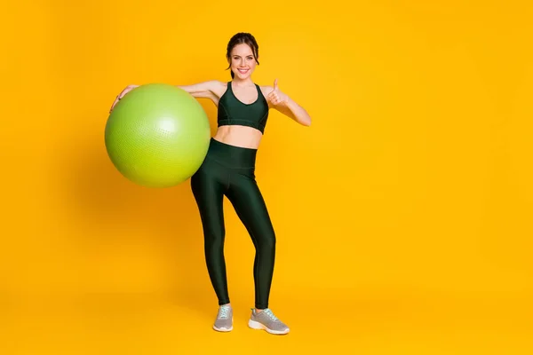 Tamanho do corpo de comprimento total vista de menina alegre agradável esportivo carregando bola de fitness mostrando polegar isolado sobre fundo de cor amarela brilhante. — Fotografia de Stock