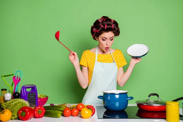 Photo amazed retro style girl prepare veggie soup boil saucepan hold ladle open cover lid impressed problem cook dish wear yellow dotted dress hair curlers isolated green color background
