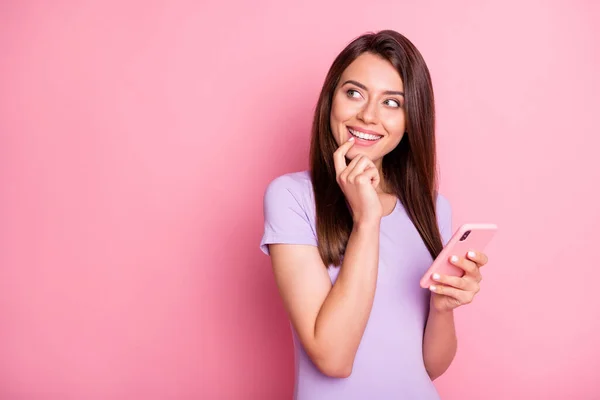 Foto de funky jovem mulher cabelo marrom segurar celular olhar espaço vazio toque queixo desgaste roxo t-shirt isolado no fundo cor-de-rosa — Fotografia de Stock