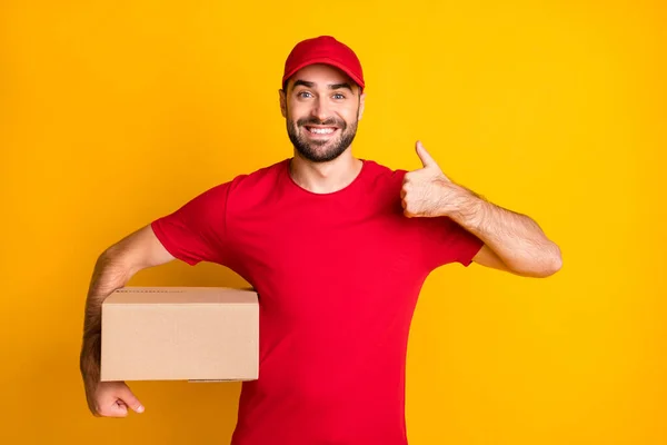 Foto di ragazzo allegro corriere vestiti di rosso consegna scatola marrone mostrando pollice su isolato sfondo di colore giallo — Foto Stock
