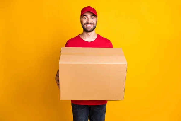 Photo of cheerful guy courier dressed red clothes delivering large brown box isolated yellow color background — Foto Stock