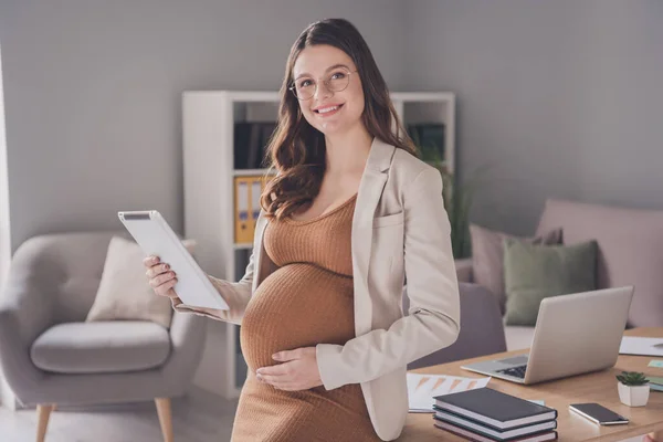 Foto portrét těhotné dámy s tabletou dotýkající se břicha sedící na stole v moderní kanceláři — Stock fotografie