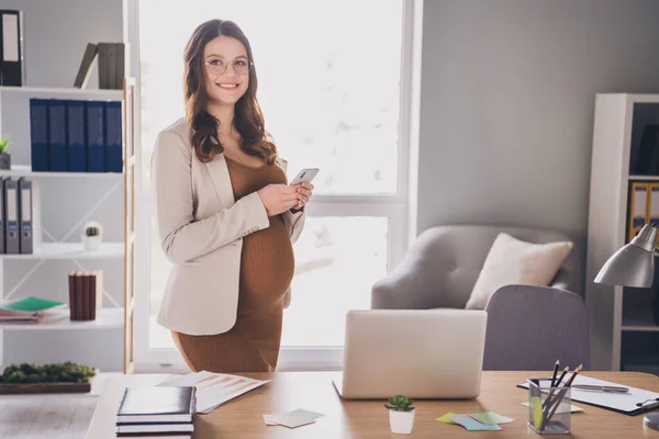 Fotografie těhotné dámy držící telefon ve dvou rukou stojící za stolem v moderní kanceláři — Stock fotografie