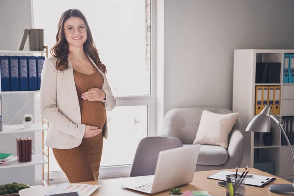 Fotografie těhotné šťastné mladé ženy ruce držet velké břicho nosit brýle oblek hnědé šaty pracovní stanice kancelář uvnitř — Stock fotografie