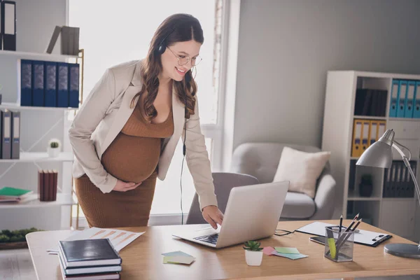 Foto portrét těhotné ženy sklánějící se k notebooku ve sluchátkách v moderní kanceláři — Stock fotografie