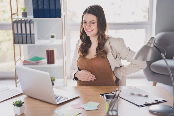 Foto portrét těhotné ženy dotýkající se páteře protahování při pohledu na notebook na stole v moderní kanceláři — Stock fotografie