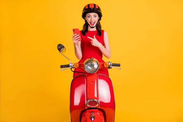 Photo of impressed young woman dressed red outfit riding vintage moped pointing modern device isolated yellow color background
