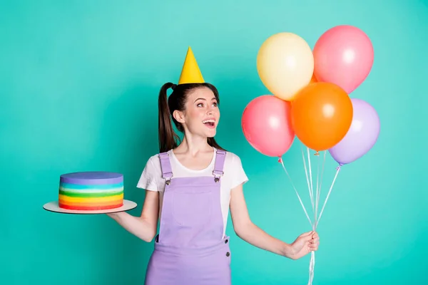 Φωτογραφία του χαριτωμένο κορίτσι κρατήσει μπαλόνια τούρτα ματιά διακόσμηση φορούν μωβ συνολικό χαρτί κώνο καπάκι απομονωμένο τυρκουάζ φόντο χρώμα — Φωτογραφία Αρχείου