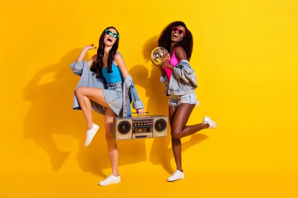 İki pozitif bayanın dansı, disko topu kasetçalar, güneş gözlüğü, kot pantolon, izole edilmiş sarı arka plan. — Stok fotoğraf