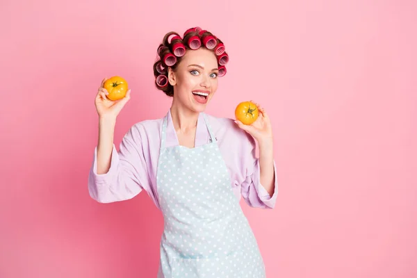 Foto van vrolijke mooie huisvrouw gekleed violet badjas rode rollen met twee gele tomaten geïsoleerde roze kleur achtergrond — Stockfoto