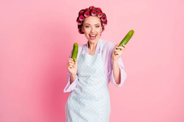 Photo of shiny dancing house wife wear dotted blue apron red rollers holding hands arms two green cucumbers isolated pink color background — Stock Photo, Image