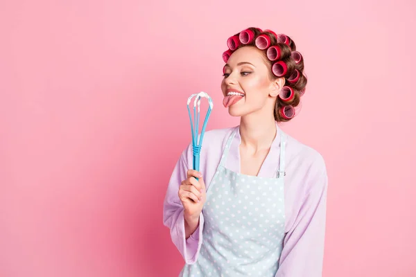 Photo of pretty house wife wear dotted blue apron red rollers looking holding licking whisk isolated pink color background — Stock Photo, Image