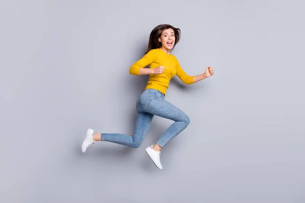 Foto de perfil em tamanho completo de hooray morena agradável senhora salto correr desgaste camisola amarela jeans tênis isolado no fundo de cor cinza — Fotografia de Stock