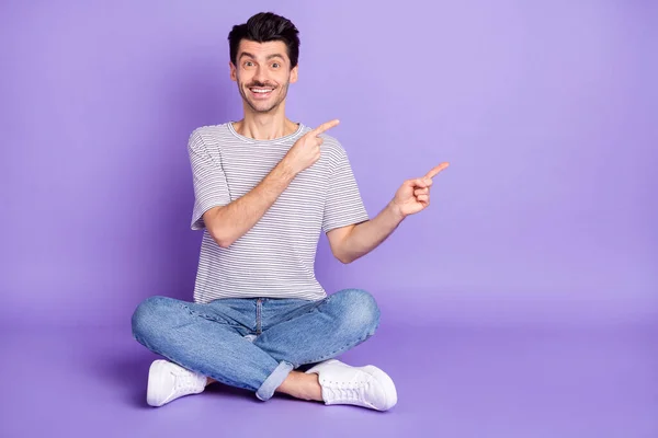 Foto di ragazzo sedersi pavimento dita dirette spazio vuoto usura strisce bianco t-shirt jeans calzature isolato sfondo viola — Foto Stock