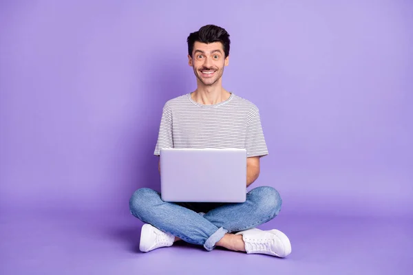 Photo of man sit floor hold pc toothy beaming smile wear black t-shirt jeans sneakers isolated purple background — Stock Photo, Image
