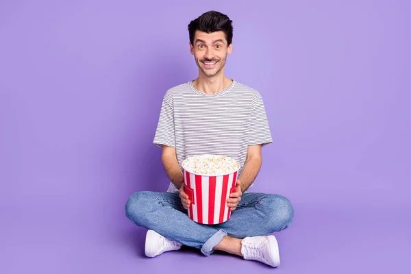 Foto di ragazzo tenere popcorn scatola dentata sorriso raggiante indossare strisce t-shirt jeans scarpe da ginnastica isolato sfondo viola — Foto Stock