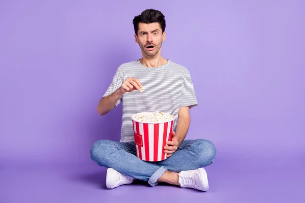 Foto di ragazzo tenere popcorn scatola spaventato bocca aperta indossare strisce bianche t-shirt jeans scarpe da ginnastica isolato sfondo viola — Foto Stock