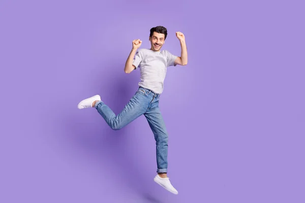 Foto av galen upphetsad kille hoppa höja nävarna bära vit randig t-shirt jeans skor isolerad violett bakgrund — Stockfoto