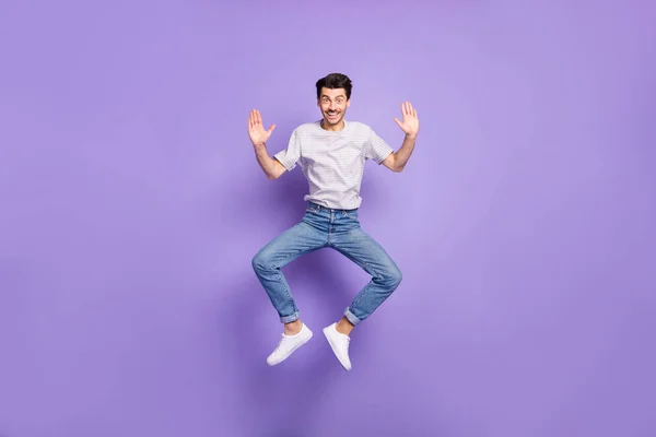 Foto di energico ragazzo attivo salto sollevare palme indossare bianco a righe t-shirt jeans calzature isolato sfondo viola — Foto Stock