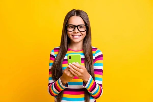 Фотопортрет веселой школьницы, печатающей на мобильнике улыбающиеся очки, изолированные на ярком желтом фоне — стоковое фото
