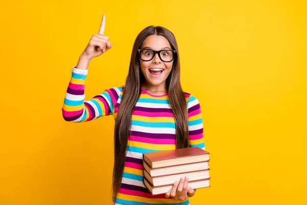 Retrato fotográfico de la colegiala sonriente feliz manteniendo libros de texto apuntando con el dedo hacia arriba idea de signo aislado color amarillo brillante fondo — Foto de Stock