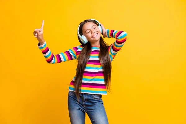 Retrato fotográfico de una niña sonriente con auriculares apuntando hacia arriba con el dedo con los ojos cerrados aislados sobre un fondo de color amarillo vivo — Foto de Stock