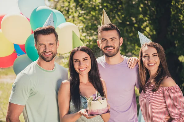 Foto de homens e mulheres alegres celebrar aniversário desgaste cone chapéus segurar bolo sorriso feliz fora no campo ao ar livre — Fotografia de Stock