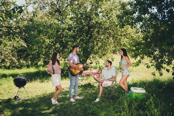 Volledige lengte foto van vier vrolijke vrienden weekend feest houden bier fles spelen gitaar chill gesprek buiten in de tuin — Stockfoto