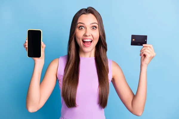 Foto porträtt av upphetsad flicka som visar mobiltelefon skärm copyspace debit plast kort isolerad på levande blå färg bakgrund — Stockfoto