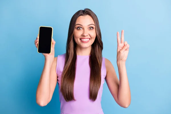Фотопортрет молодої дівчини, що демонструє сенсорний екран мобільного телефону, що показує v-знак ізольовано на яскравому синьому кольоровому фоні — стокове фото
