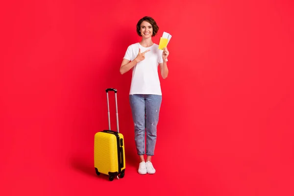 Вид в полный рост на симпатичную жизнерадостную девушку, держащую в руках билеты на экскурсию, изолированную на ярко-красном фоне — стоковое фото