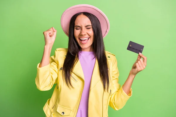Fénykép funky meglepett lány hold hitelkártya emelés ököl csukott szemek kopás sárga kabát rózsaszín kalap elszigetelt zöld háttér — Stock Fotó